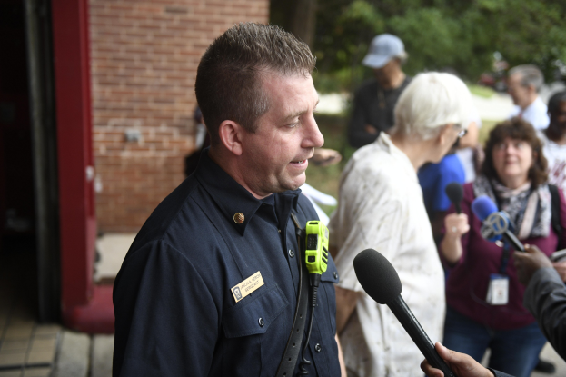 Polizeibeamter in Uniform, der zu einer Gruppe von Menschen spricht, mit Mikrofonen, Bäumen und einem Gebäude im Hintergrund.