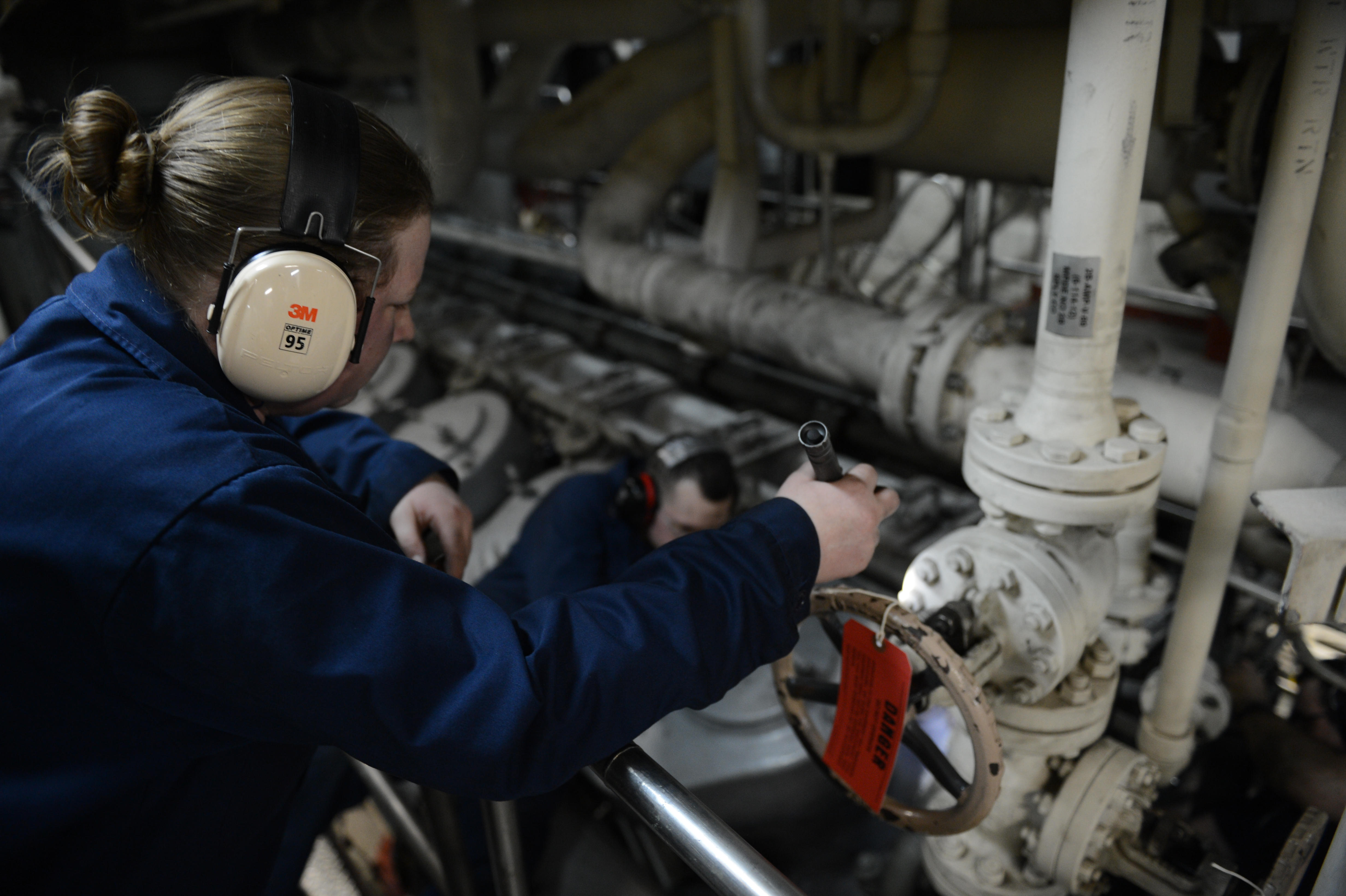 Zwei Frauen in blauen Uniformen arbeiten an einer Maschine in einer Öl- und Gasindustrieumgebung, eine hält ein Objekt und trägt Kopfhörer, mit Rohren im Hintergrund.