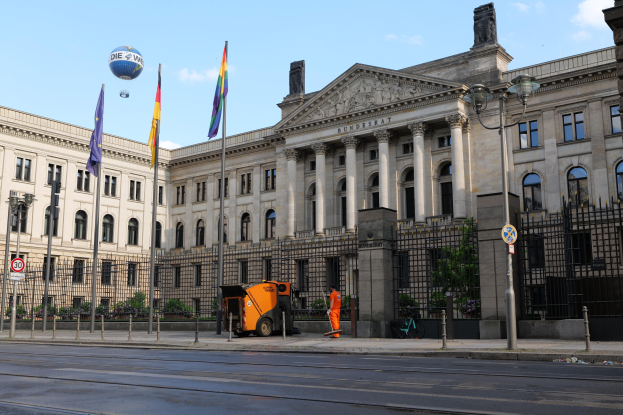 Großes modernes Regierungsgebäude mit vielen Fenstern, umgeben von Straßenmöbeln, Fahrzeugen, Fußgängern und Bäumen, vor einem bewölkten Himmel.