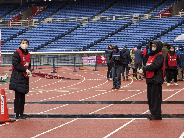 Gruppe von Menschen auf einer Rennstrecke, die ein Band halten, mit einem Verkehrskegel links und Event-Infrastruktur im Hintergrund.