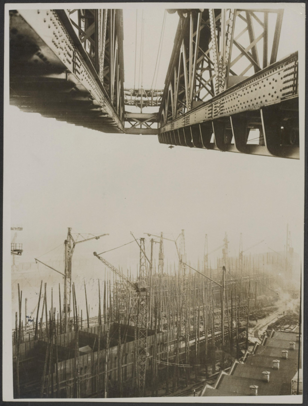 Schwarz-weiß-Foto einer Baustelle mit einer teilweise gebauten Brücke im Hintergrund, mit Pfählen und Kränen unten.