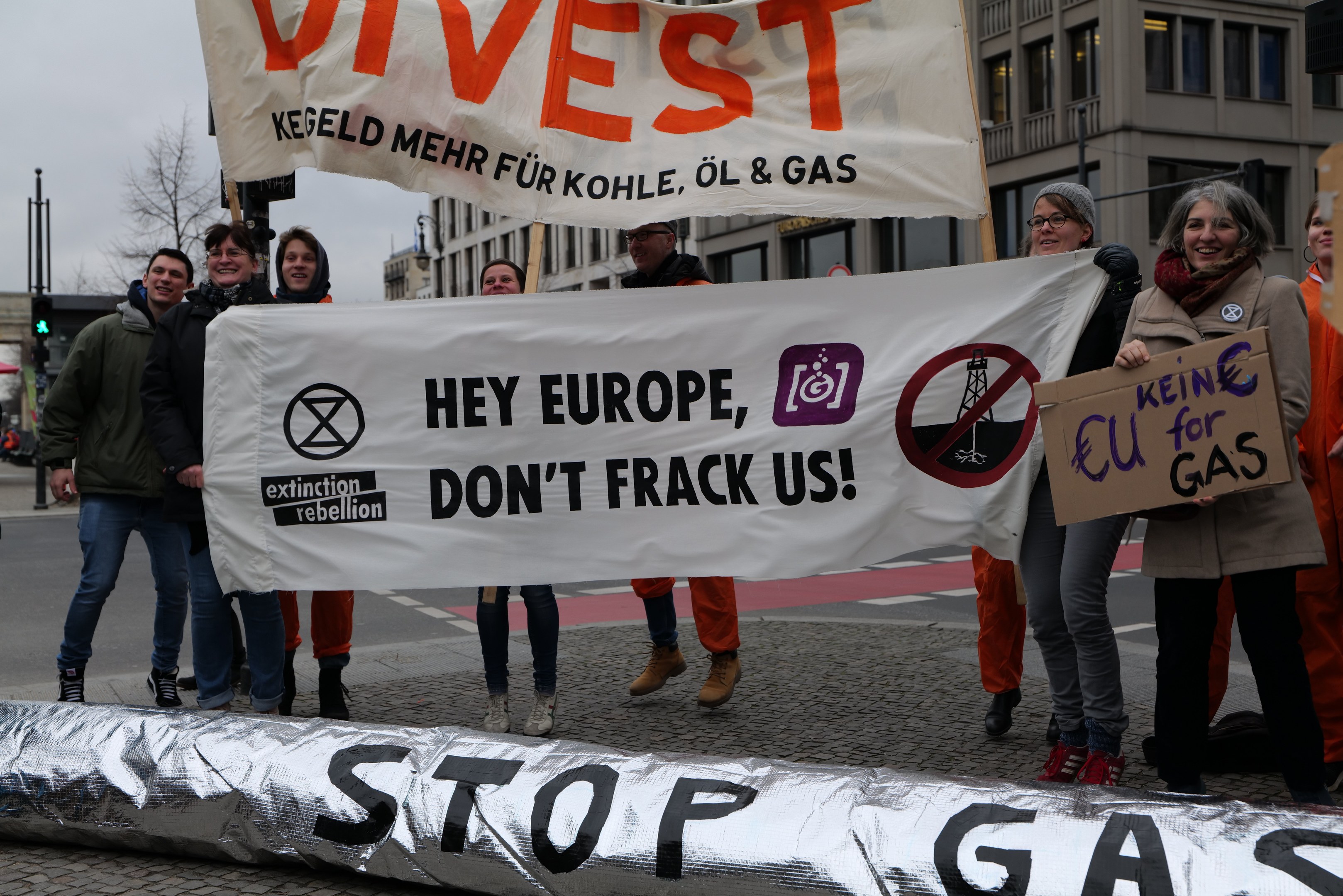 Gruppe von Menschen auf einer Straße mit einem "Stoppt Gas"-Schild, die gegen die Europäische Union protestieren, mit Gebäuden, Bäumen, Polen, Verkehrszeichen und einem klaren blauen Himmel im Hintergrund.
