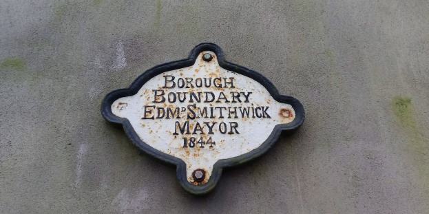 Ein Metallschild an einem Gebäude mit schwarzem Hintergrund und weißer, fetter, zentrierter Schrift, die 'Borough Boundary Edm Smithwick Mayor' lautet.