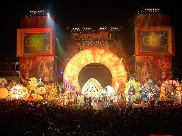 Eine große Menschenmenge bei Nacht vor einer Bühne mit einem Bildschirm, auf dem "Carnaval de Vinaros" und farbige Beleuchtung zu sehen ist.