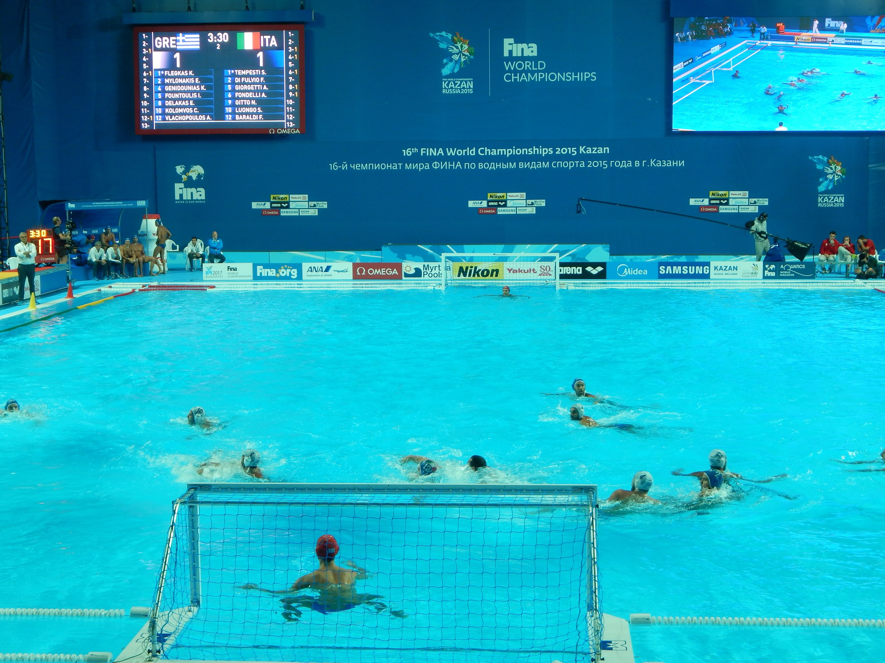 Wasserballspieler bei den FINA-Weltmeisterschaften 2016 in Kazan im Pool gegeneinander antretend, mit Zuschauern auf der linken Seite und einer Anzeigetafel mit Event-Branding im Hintergrund.