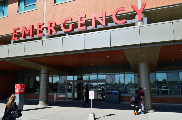 Gruppe von Menschen, die vor einem Notaufnahme-Gebäude mit Glaswänden, Säulen und einem Namensschild gehen, neben Schildern und einem Müllcontainer.