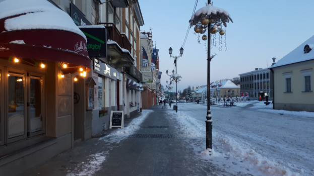 Eine schneebedeckte Stadtstraße mit Gebäuden auf beiden Seiten, beleuchtet von Straßenlaternen, mit Menschen, die auf den Gehwegen gehen und Bäumen im Hintergrund bei klarem Himmel.