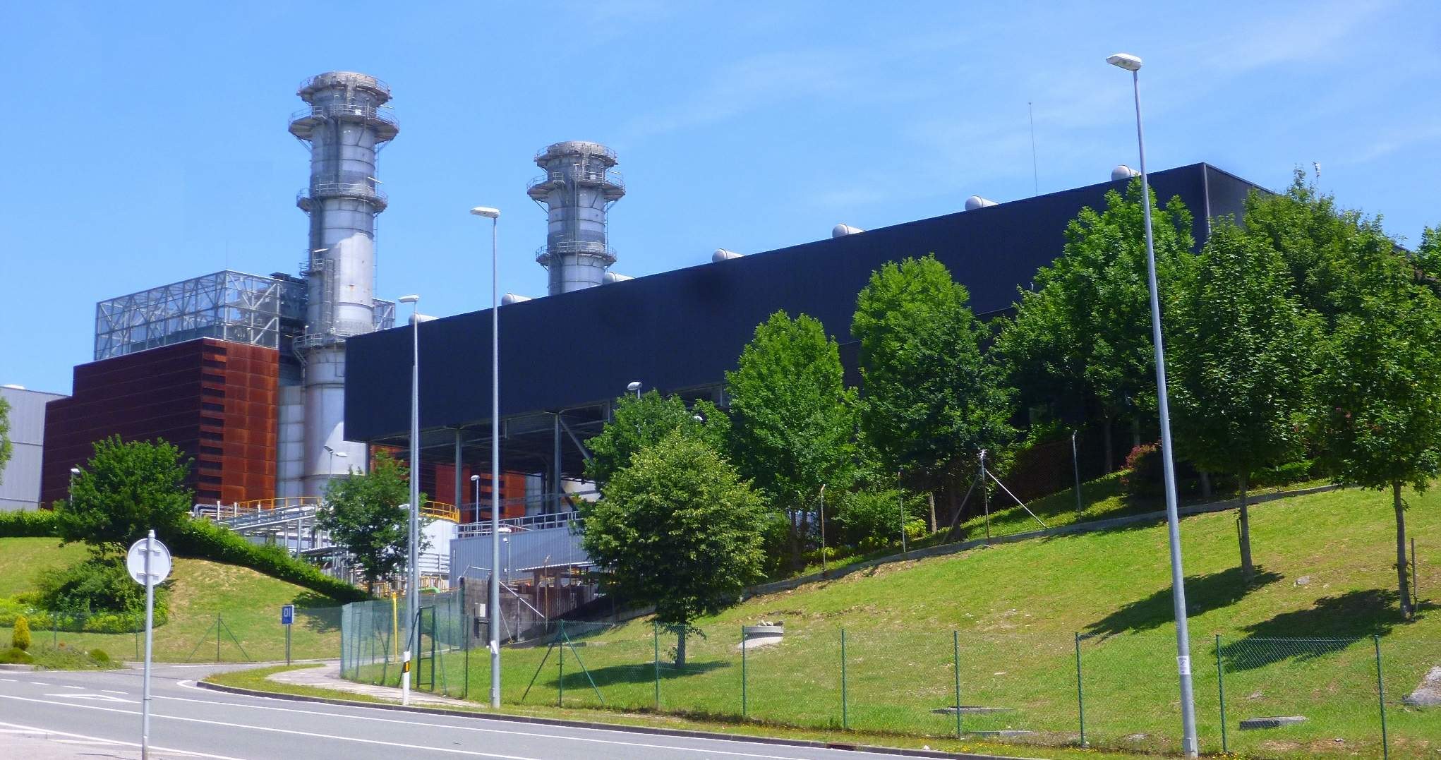 Großes Industriegebäude mit zahlreichen Fenstern, als Kraftwerk identifiziert, umgeben von städtischer Infrastruktur einschließlich Straßenlaternen, Fahrzeugen und Grünflächen unter einem bewölkten Himmel.