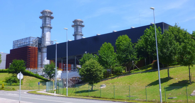 Großes Industriegebäude mit zahlreichen Fenstern, als Kraftwerk identifiziert, umgeben von städtischer Infrastruktur einschließlich Straßenlaternen, Fahrzeugen und Grünflächen unter einem bewölkten Himmel.