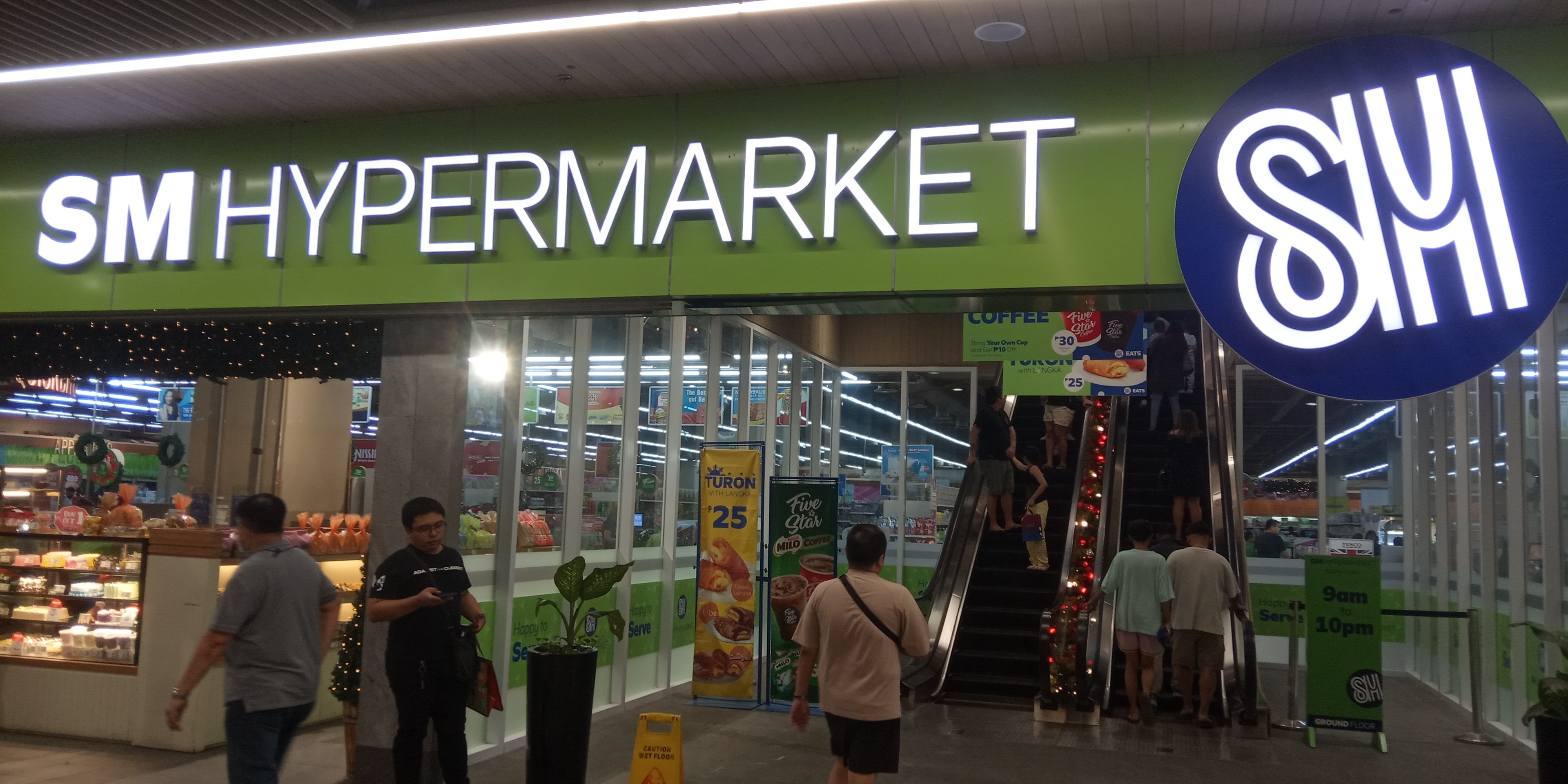 Group of people walking in front of a brightly lit "SM Hypermarket" store with ceiling lights, text boards, and houseplants visible.