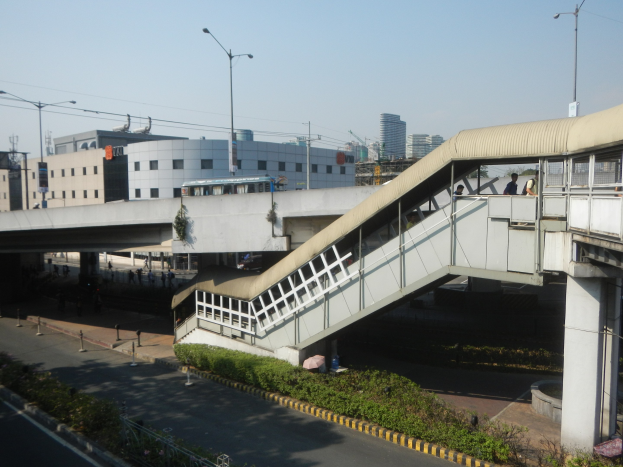 Fußgängerbrücke über einer Straße mit Menschen, umgeben von Gebäuden, Straßeninfrastruktur, Fahrzeugen und Grünflächen unter einem klaren blauen Himmel.