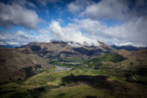Ein atemberaubender Blick von der Spitze eines Berges in Queenstown, Neuseeland, der üppiges grünes Gras, Bäume, eine kurvenreiche Straße und einen Himmel mit weißen, flauschigen Wolken zeigt.