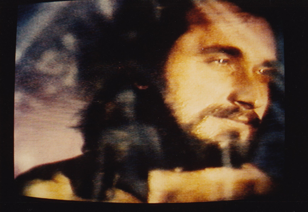 Ein Mann mit langem dunklem Bart und Haar, der ein weißes Hemd trägt, erscheint auf einem Fernsehbildschirm mit einem ernsten Gesichtsausdruck und schaut direkt in die Kamera.