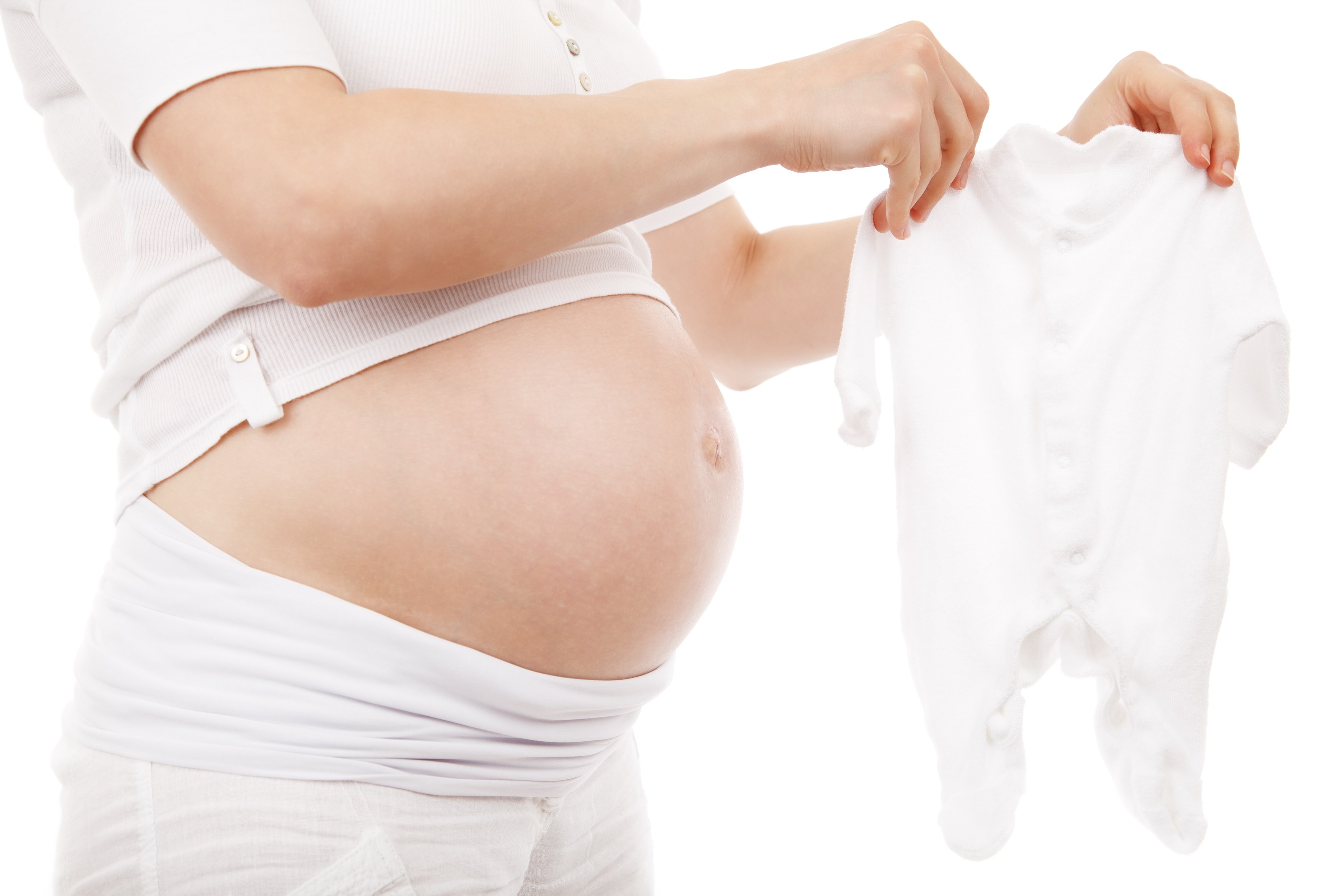 Eine schwangere Frau in einem weißen Kleid hält ein weißes Baby-Bodysuit, strahlt glücklich.