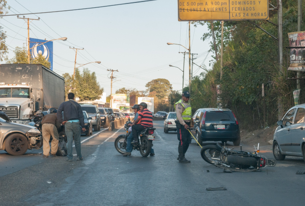 Eine Gruppe von Menschen steht in der Nähe eines verunglückten Motorrads an der Straße mit mehreren Fahrzeugen, Bäumen, Strommästen und einem klaren Himmel im Hintergrund.