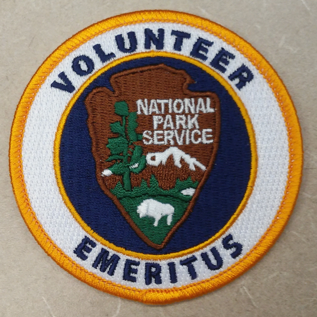 Ein rundes Patch mit einem blauen Rand, weißem Hintergrund und fetter schwarzer Schrift, die den Text 'Volunteer National Park Service Emeritus' in der Mitte eines gelben Kreises zeigt.