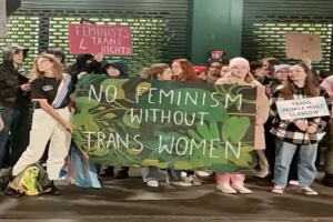 Eine Gruppe von Menschen hält ein Transparent mit der Aufschrift "Kein Feminismus ohne Transfrauen" und Schilder in einem öffentlichen Raum mit einer Wand und einem Rollladen im Hintergrund.
