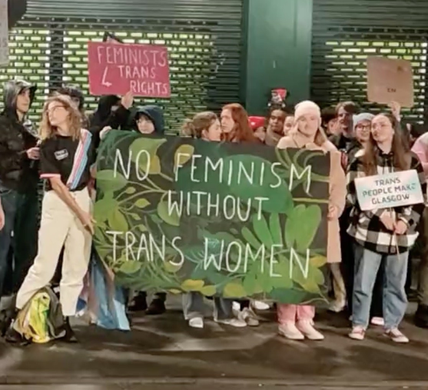 Eine Gruppe von Menschen hält ein Transparent mit der Aufschrift "Kein Feminismus ohne Transfrauen" und Schilder in einem öffentlichen Raum mit einer Wand und einem Rollladen im Hintergrund.