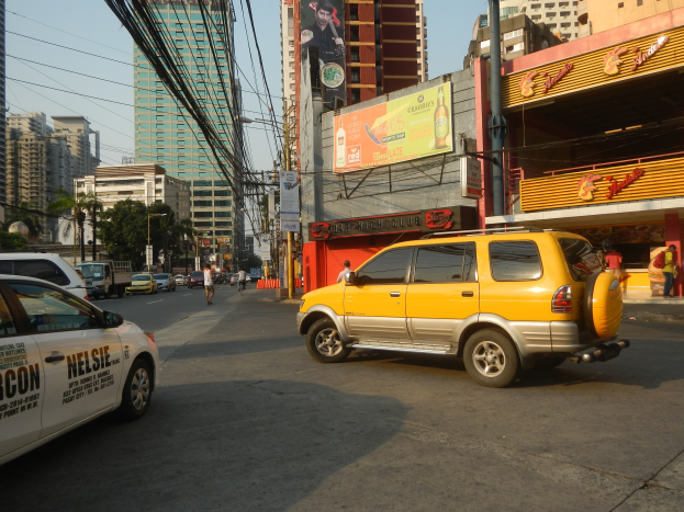 Ein gelbes Taxi fahrt eine Stra├če mit hohen Geb├Ąuden entlang, Menschen auf dem Gehweg, Strommasten mit Kabeln und B├Ąumen.