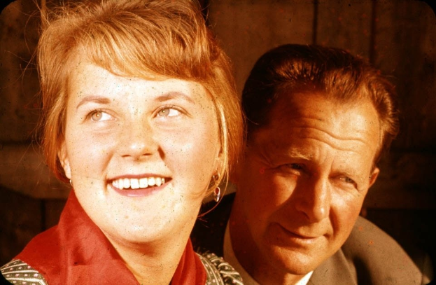 Zwei Menschen in inniger Umarmung, wobei der Mann seinen Arm um die Taille der Frau gelegt hat und ihr Kopf auf seiner Schulter ruht, vor einer schmucklosen Wand stehend.