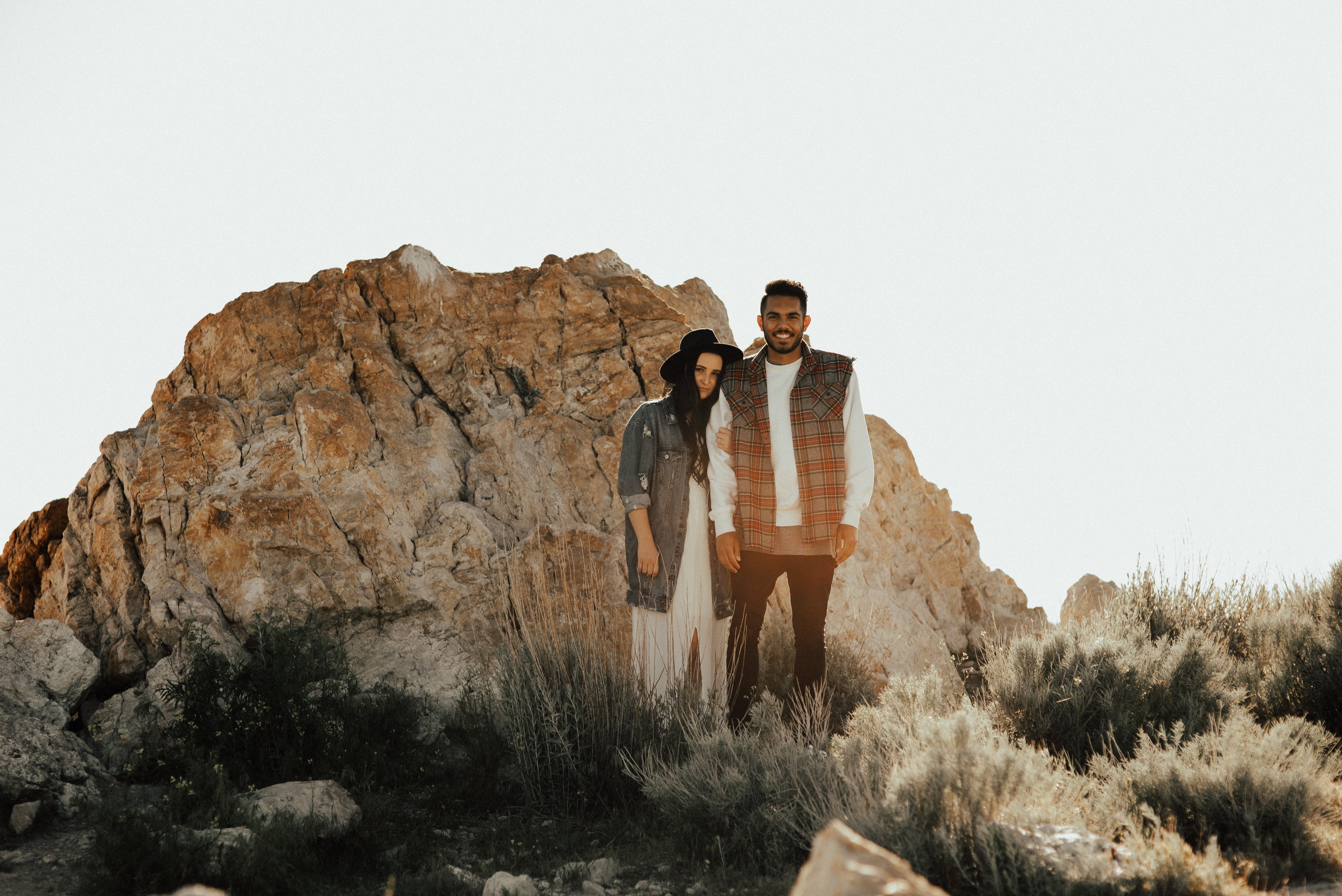 Ein Mann und eine Frau stehen lachend auf einem Felsen in der Wüste, umgeben von grünem Gras und Pflanzen unter einem klaren blauen Himmel während einer Fotosession zur Verlobung.