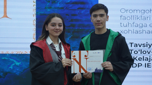 Ein Mann und eine Frau in Graduationsroben stehen nebeneinander, jeder hält ein Zertifikat in der Hand, mit einem Bildschirm im Hintergrund.