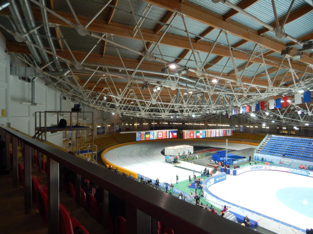 Großes Innen-Eisstadion mit zahlreichen Menschen auf dem Eis, umgeben von Sitzplätzen, Geländern, Brettern, Leuchten und strukturellen Elementen, mit einer Wand im Hintergrund.
