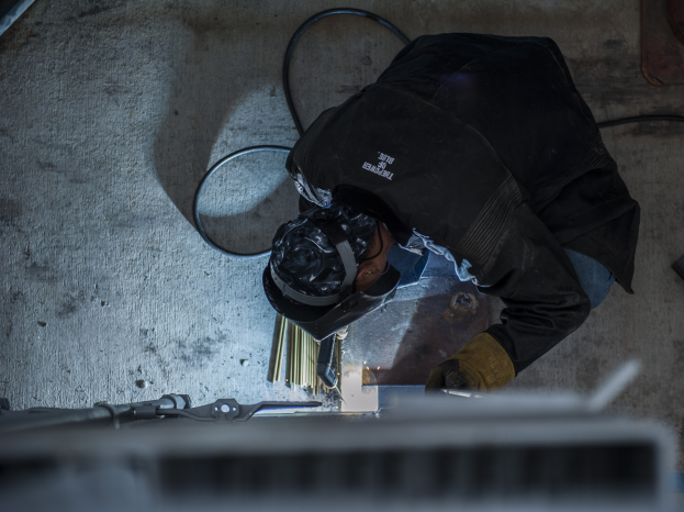 Ein Schweißer in Schutzausrüstung bedient eine Schweißmaschine an einem Metallstück, mit Werkzeugen und Gegenständen auf dem Boden verstreut.