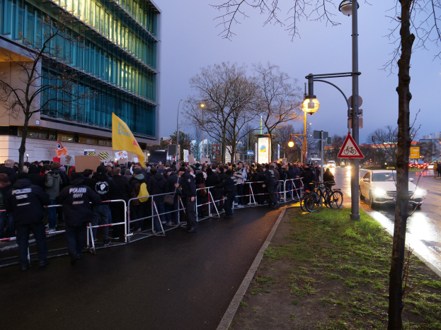 Eine große Gruppe von Menschen steht vor einem Gebäude und hält Schilder und Protestplakate, mit Barrikaden, Fahrrädern, Laternenmasten, Schautafeln, Bäumen und Gras auf dem Boden in Berlin.