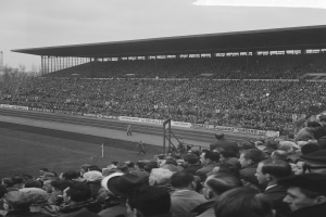 Schwarzes und weißes Foto eines vollen Fußballstadions mit Zuschauern, die ein Spiel verfolgen, mit Bannern, Pfosten, einem Schuppen, Bäumen, einem Turm und einem bewölkten Himmel.