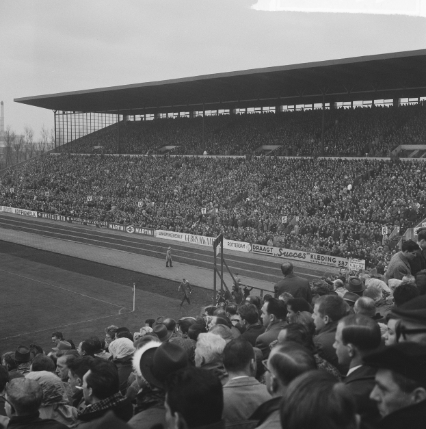 Schwarzes und weißes Foto eines vollen Fußballstadions mit Zuschauern, die ein Spiel verfolgen, mit Bannern, Pfosten, einem Schuppen, Bäumen, einem Turm und einem bewölkten Himmel.