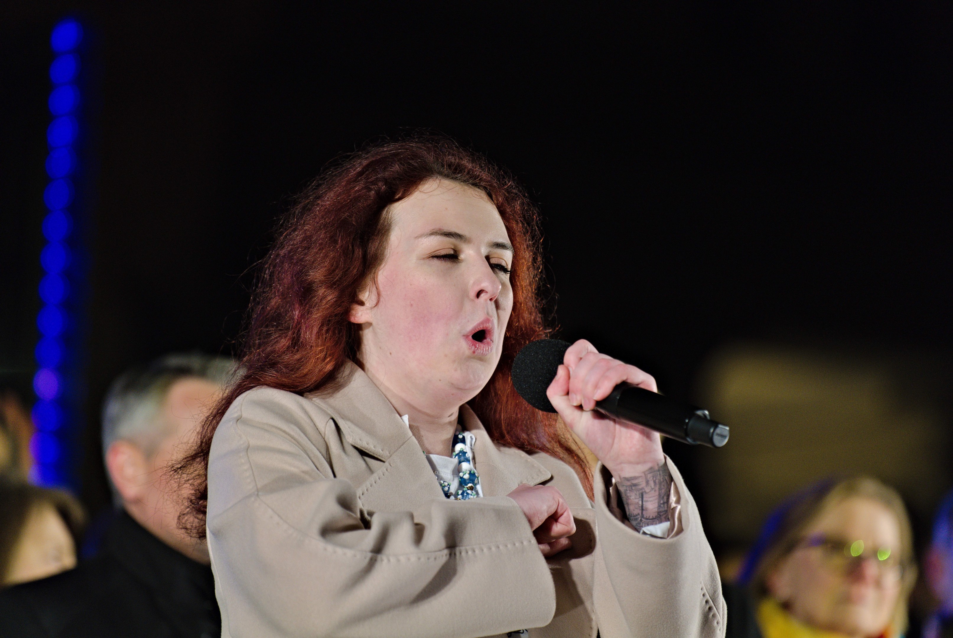 Eine Frau singt in ein Mikrofon auf der Bühne mit zwei Personen dahinter, unter Bühnenbeleuchtung.