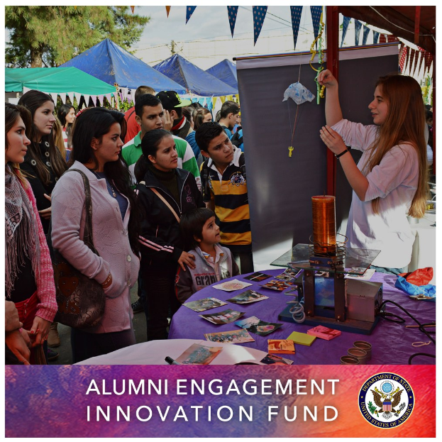 Gruppe von Menschen um einen Tisch versammelt, auf dem ein Schild für den "Alumni Engagement Innovation Fund" steht, mit verschiedenen Gegenständen darauf, vor einer Kulisse aus Zelten, Bäumen und einem klaren blauen Himmel, einschließlich Text und Logo unten.