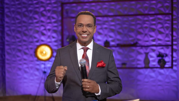 Stephen Colbert, Moderator von *The Daily Show with Trevor Noah*, steht auf einer Bühne und hält ein Mikrofon, trägt einen Anzug, Krawatte und Brille, mit einer Lampe und einem Ständer dahinter.