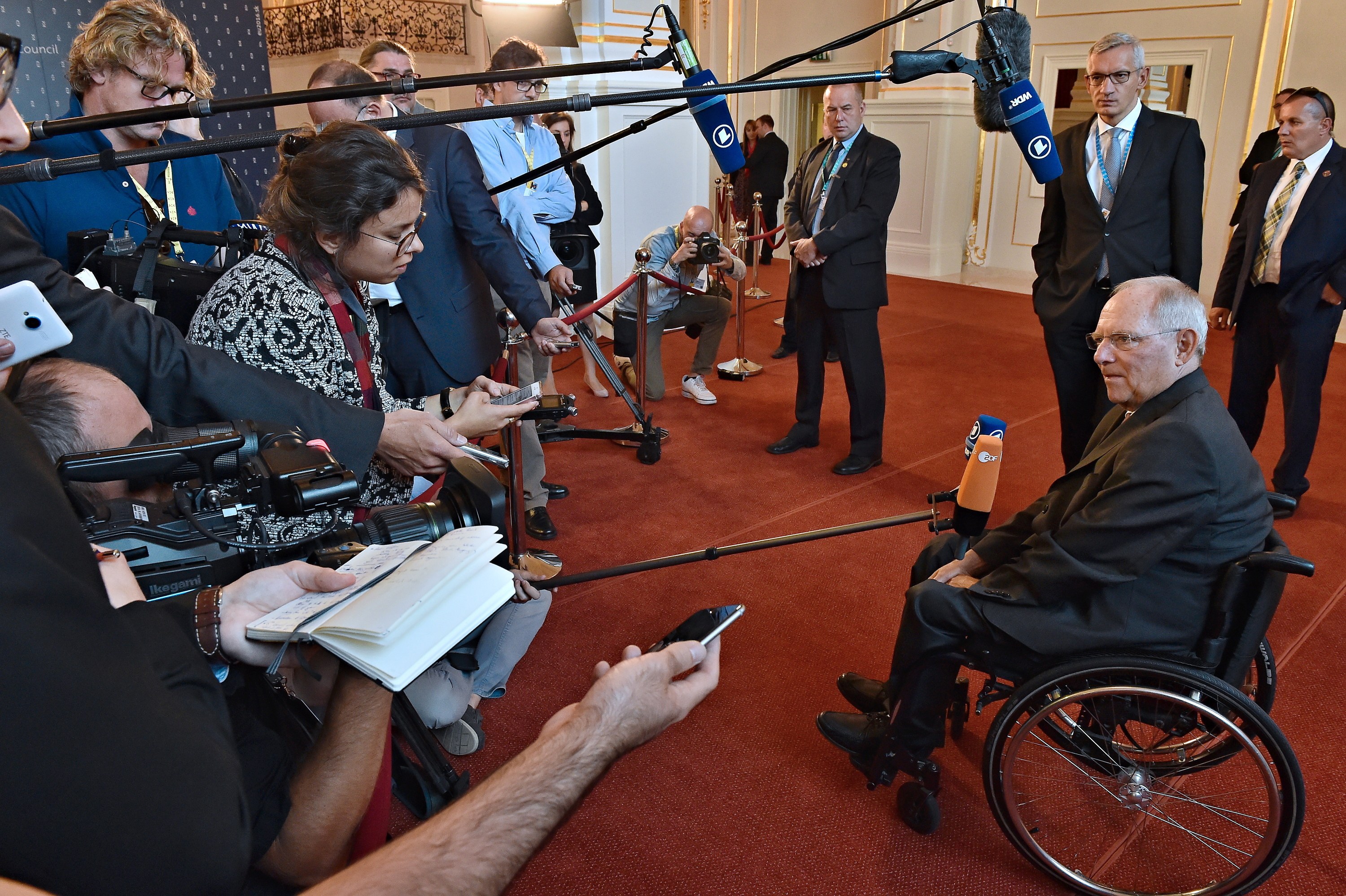 Ein Mann im Rollstuhl umgeben von Reportern mit Kameras, Handys und Mikrofonen, mit einem Banner, Scheinwerfern und einer Wand im Hintergrund.