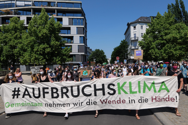 Gruppe von Menschen in Masken, die ein Banner mit der Aufschrift "Aufbruchsklima" vor einem Gebäude mit Bäumen und wolkenlosem Himmel halten.