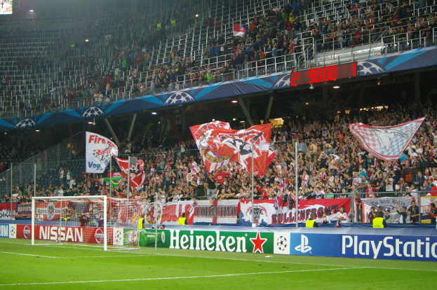 Ein Fuballspiel in einem hell erleuchteten Stadion mit einer grossen Zuschauermenge, einem Torpfosten in der Mitte, Bannern drumherum und einem Bildschirm oben links.