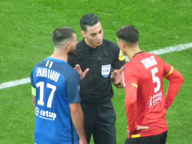 Schiedsrichter spricht mit zwei Männern auf einem Fußballfeld, einer in Schwarz und der andere in einem blauen und roten T-Shirt mit Text, beide tragen eine Brille.