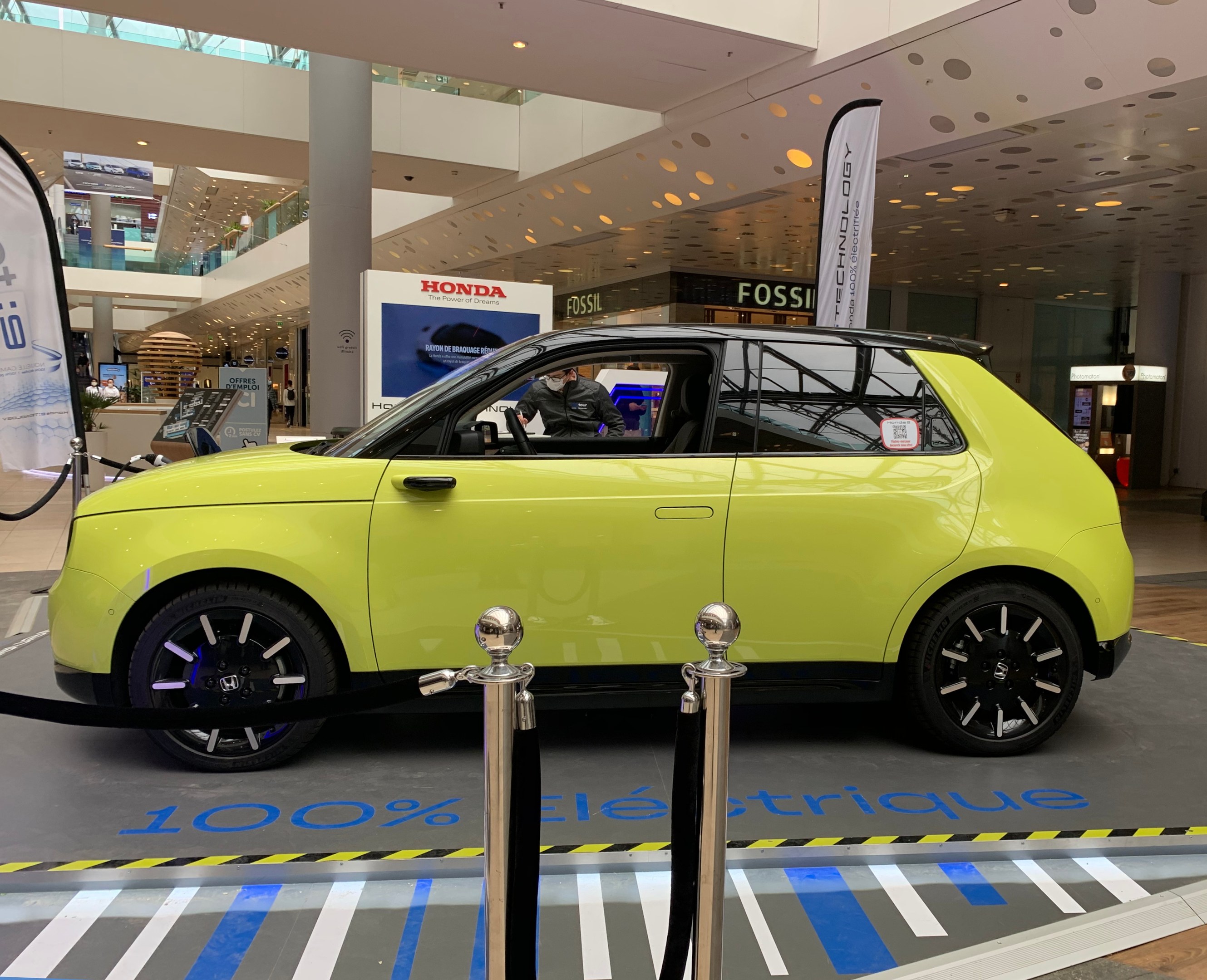 Ein gelbes wasserstoffbetriebenes elektrisches Auto vor einem Gebäude mit Absperrpollern, Bannern, Menschen, Schildern, einer Topfpflanze, Deckenleuchten und Ventilatoren.