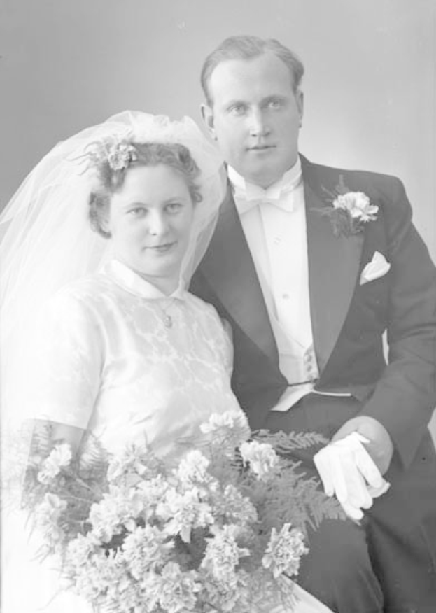 Ein Schwarz-Weiß-Foto eines strahlenden Mannes und einer Frau an ihrem Hochzeitstag, wobei die Frau einen Blumenstrauß hält und der Mann einen Anzug trägt und liebevoll in die Augen schaut.