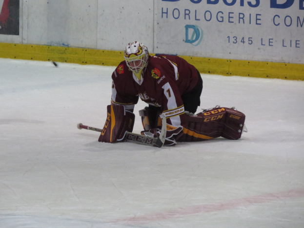 Eishockey-Spieler in rot-gelber Uniform bei einem Save, mit einer Wand und Text im Hintergrund.