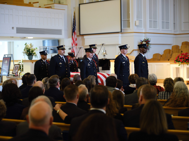 Eine Gruppe uniformierter Personen mit Mützen steht vor sitzenden Trauergästen bei einer Beerdigung, mit Blumenarrangements, einer Fahne, einem Bildschirm und einer Wand im Hintergrund.