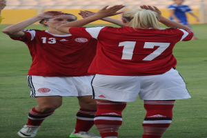 Zwei Frauen in roten T-Shirts und weißen Shorts stehen auf einem Fußballfeld und klatschen begeistert in die Hände, im Hintergrund ist eine Person in einem blauen Kleid, eine gelbe Tafel und einige Stühle zu sehen.