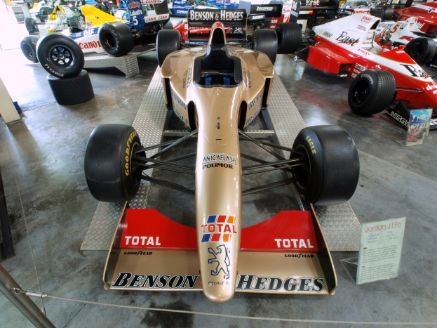 Rennwagen in einem Motorsportmuseum ausgestellt, getrennt von Zuschauern durch einen Metallzaun, mit informativen Texten im Hintergrund.