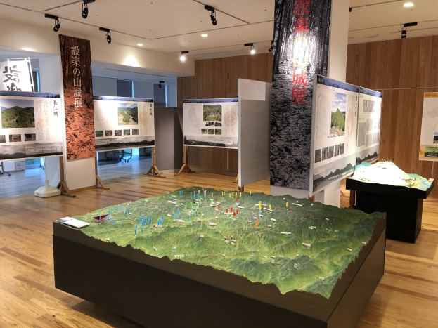 Ausstellungsraum mit verschiedenen historischen Karten Japans, darunter Wandkarten, ein Tisch mit einer Karte, Banner, Informationsschilder auf Ständern, Deckenleuchten und eine hölzerne Rückwand.