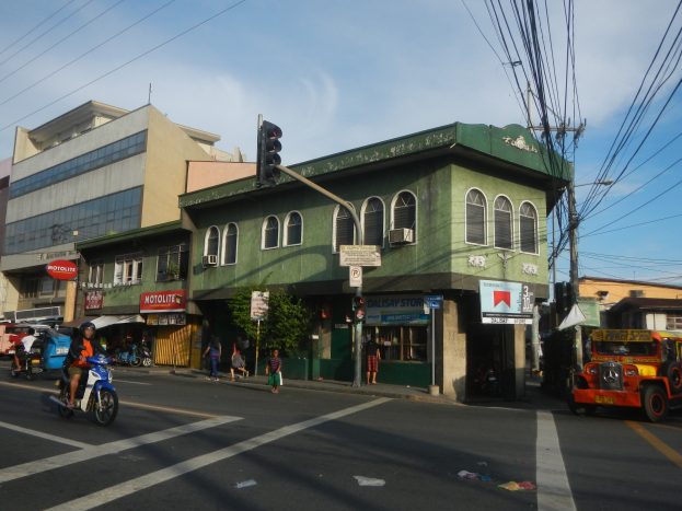 Eine belebte Stadtstraße mit einem grünen Gebäude im Hintergrund, Fahrzeugen, Fußgängern, einer Ampel, einem bewölkten Himmel und Oberleitungen.