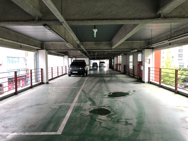 Ein mehrgeschossiges Parkhaus mit geparkten Autos, Gel├Ąndern, Pfeilern, Deckenleuchten, freiliegenden Rohren und umgebenden B├Ąumen, vor einem Hintergrund von Geb├Ąuden mit Fenstern in einer Stadtlandschaft.