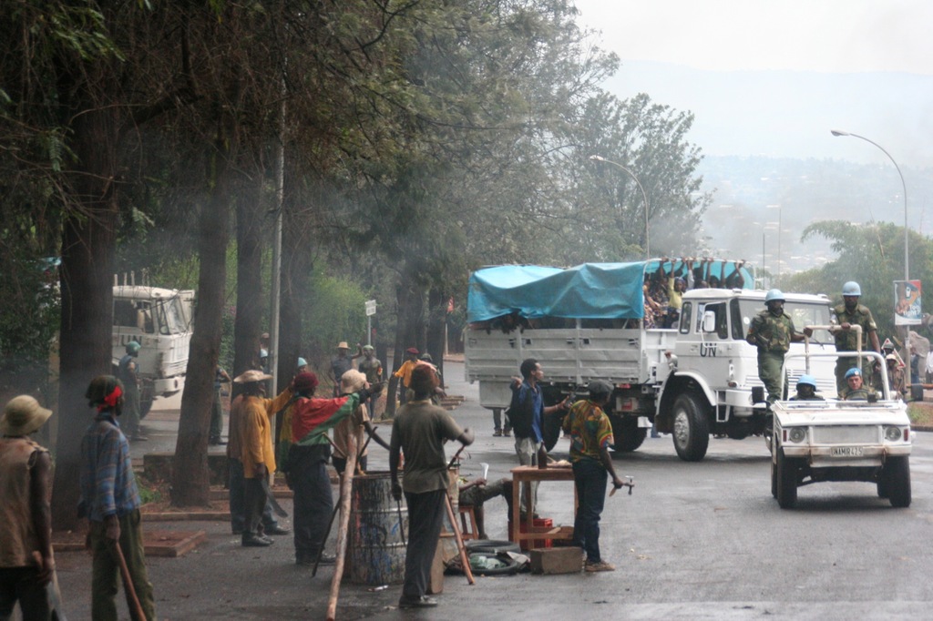 Arbeiter f√ľhren Aufgaben neben einer Stra√üe mit einem Lastwagen durch, auf dem eine gro√üe Menge Menschen steht, w√§hrend Beamte in einem Fahrzeug in der N√§he fahren, umgeben von B√§umen und Stra√üenlaternen.