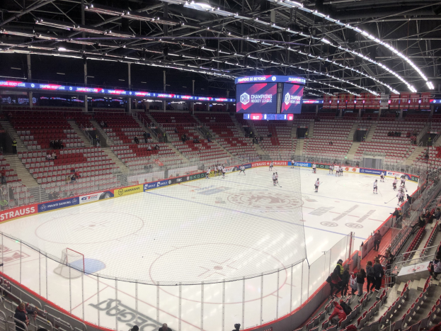 Eishockeyspiel in einer großen Arena mit Zuschauern, eine Eisfläche umgeben von einem Zaun, ein Display, das "St. Louis Blues vs Detroit Red Wings" anzeigt, Metallrahmen, Deckenleuchten und ein Dach.