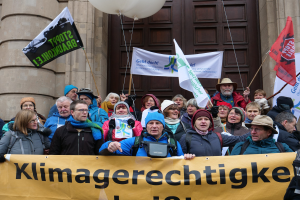 Eine Gruppe von Menschen steht vor einem Gebäude mit Säulen und einer Tür, hält ein Transparent mit Text, Fahnen mit Stangen und Luftballons während eines Klimastreiks in Deutschland.
